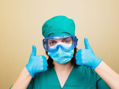 Female Doctor with thumbs up in green suit and blue mask gloves and protective glasses on yellow background. Selective focus. 2BDJK1N Female Doctor with thumbs up in green suit and blue mask gloves and protective glasses on yellow background. Selective focus.